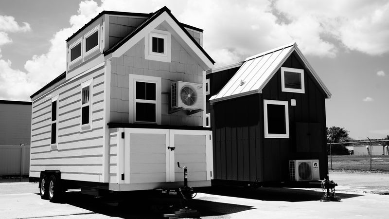 Black-and-white photo of two tiny houses on wheels: a white one with a loft dormer and mini-split, and a dark board-and-batten one with a metal roof